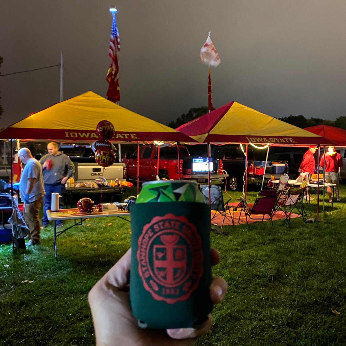 Person holding a can cooler with a logo at an outdoor event with tents and people.