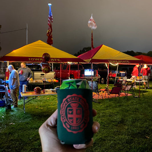 Person holding a can cooler with a logo at an outdoor event with tents and people.
