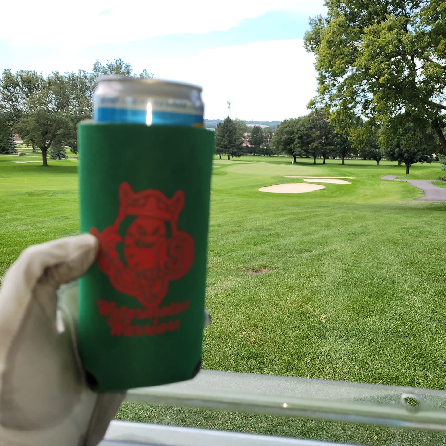 Hand holding a can with a green sleeve featuring a red logo in a park setting