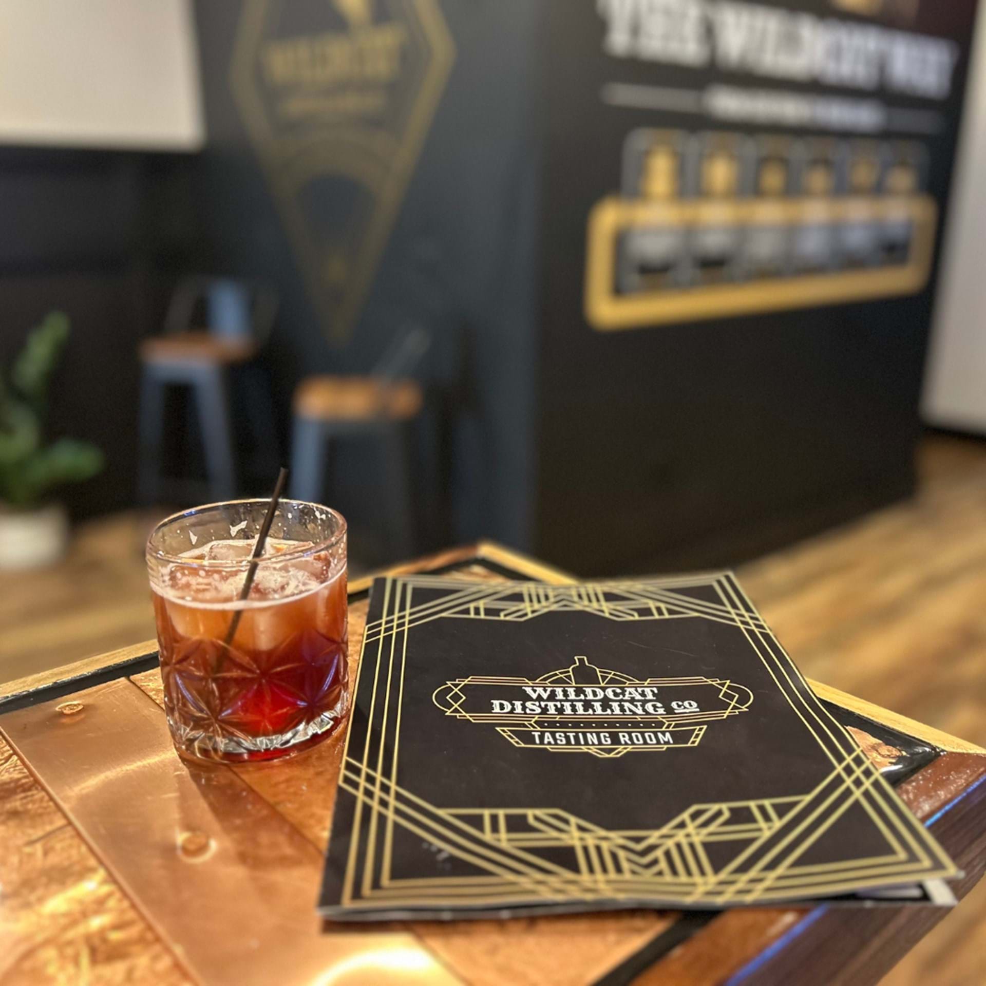 Glass of a red cocktail with a straw on a coaster next to a 'Wildcat Distilling Co. Tasting Room' menu.